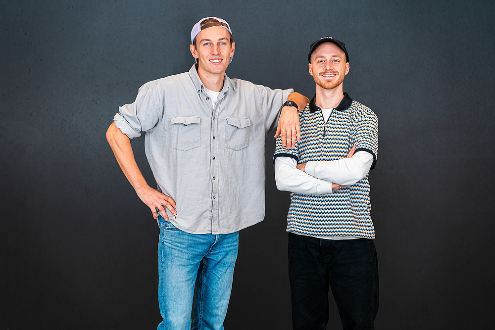 Es stehen zwei Männer im Alter von 20-30 Jahren nebeneinander. Der linke Mann trägt eine Kappe falsch herum, ein graues lockeres Hemd und eine hellblaue Jeans. Er lächelt in die Kamera und legt seinen linken Arm cool auf die Schulter des Mannes, der neben ihm steht. Neben ihm steht ein junger Mann, der ein schwarz-weiß gestreiftes Oberteil mit schwarzem Kragen trägt. Er hat eine schwarze Hose und eine schwarze Kappe an und lächelt fröhlich in die Kamera.