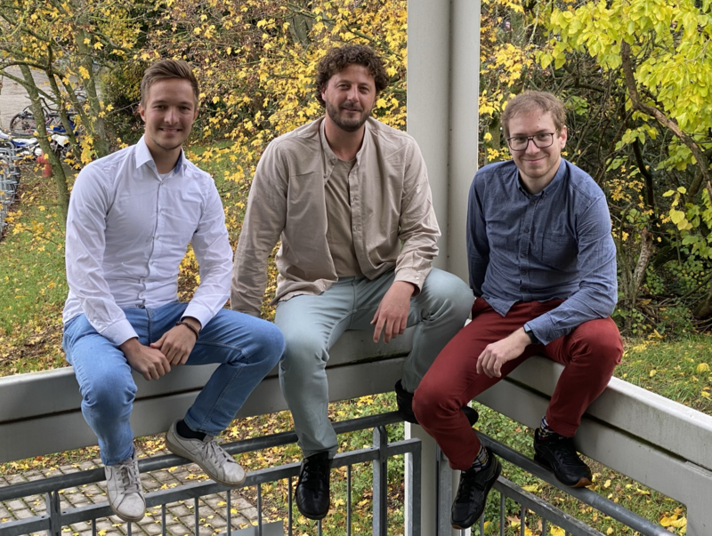 Auf dem Bild sind drei Männer zwischen 20 und 30 Jahren zu erkennen. Sie sitzen lässig auf einem Geländer und lachen in die Kamera. Der Mann links trägt ein weißes Hemd, eine hellblaue Jeans und weiße Sneaker. Er hat braune Haare. Der in der Mitte sitzende Mann trägt ein beigefarbenes Hemd, eine graue Hose und schwarze Schuhe. Er hat braune Haare und einen braunen Bart. Rechts sitzt ein blondhaariger Mann mit Brille, der ein blaues Hemd, eine rote Hose und schwarze Schuhe trägt..