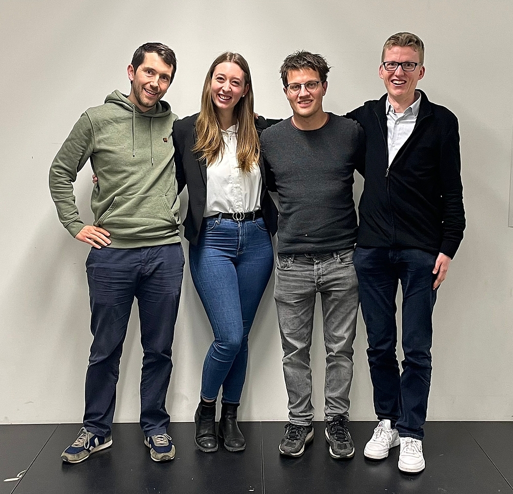 Auf dem Bild schauen vier Personen lächelnd in die Kamera. Links steht ein Mann in den 20ern, der einen Pullover in der Farbe Khaki, eine dunkle Hose sowie schwarz-weiße Sneaker trägt. Er hat braune Haare. Rechts neben ihm steht eine junge Frau in den 20ern. Sie trägt einen dunklen Blazer mit einer weißen Bluse, einer blauen Jeans und schwarzen Stiefeletten. Sie hat dunkelblond-hellbraunes Haar. Rechts neben der Frau befindet sich ein junger Mann in den 20ern. Er hat braune Haare, trägt einen dunkelgrauen Pullover, eine hellgraue Jeans und dunkle Sneaker. Ganz rechts befindet sich ein blonder junger Mann mit Brille, der eine schwarze Jacke mit einem weißen Hemd darunter sowie eine dunkle Hose und weiße Sneaker trägt.