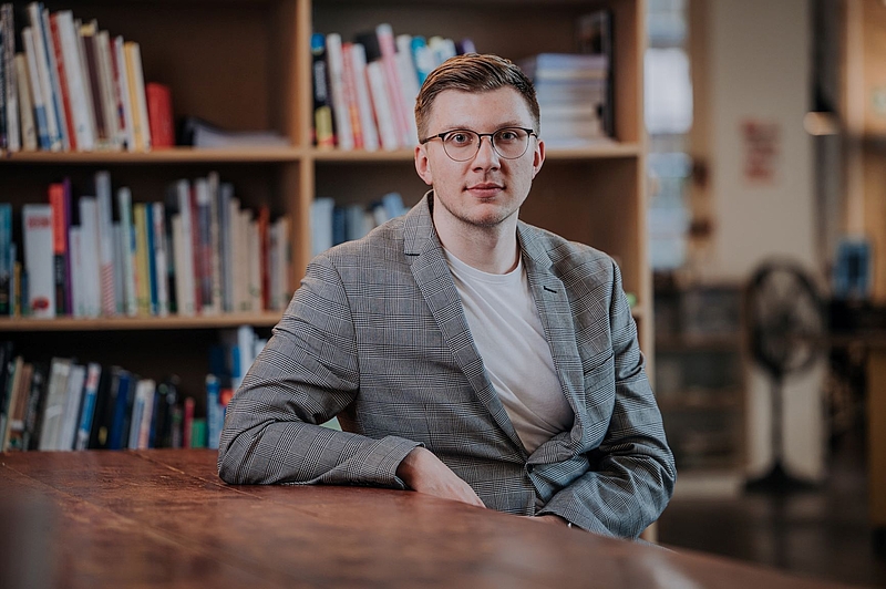 Auf dem Bild erkennt man einen Mann in den 20ern, der einen mittelgrauen Blazer und ein beigefarbenes Oberteil trägt. Der Mann hat hellbraune Haare und trägt eine Brille. Er sitzt vor einem Bücherregal.