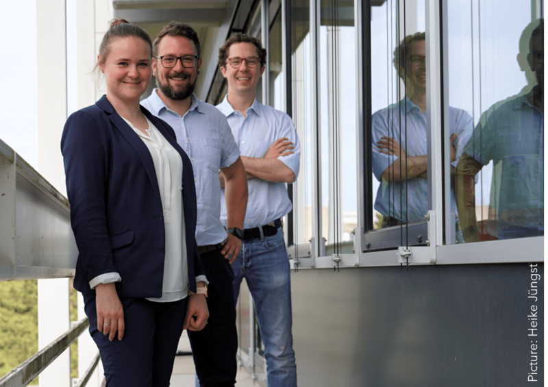 Drei Personen sind auf dem Bild zu erkennen. Sie stehen auf einem balkonähnlichen Bereich vor Fenstern. Links ist eine Frau, die einen dunkelblauen Blazer und ein weißes Oberteil trägt. Sie ist in den 20ern, trägt einen Dutt und lächelt in die Kamera. In der Mitte steht ein Mann mit Bart und Brille, der sich in den 30ern befindet. Er trägt ein hellblaues Hemd und eine dunkle Hose. Neben ihm steht ein jüngerer Mann, der die Arme ineinander verschränkt und in die Kamera lacht. Er trägt eine Brille und hat leichtet Locken.